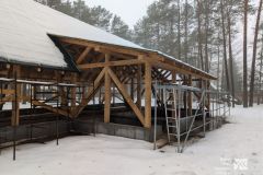 Drewniana konstrukcja budynku w trakcie budowy. Otoczenie pokryte śniegiem. W tle las.