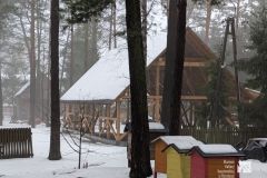 Przed drewnianą konstrukcją budynku na etapie budowy stoją trzy ule, w tle las. Otoczenie pokryte śniegiem.