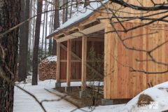 Drewniana konstrukcja budynku na etapie budowy, wokół sosnowy las. Za konstrukcją sterta porąbanego drewna, okolica pokryta śniegiem.
