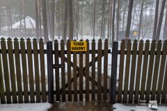 Niskie drewniane ogrodzenie, na którym widnieje tabliczka z napisem teren budowy, w tle las i drewniany maneż. Okolica pokryta śniegiem.