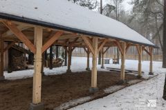 Drewniana konstrukcja, za nią leży porąbane drewno. Wszystko pokryte jest śniegiem.