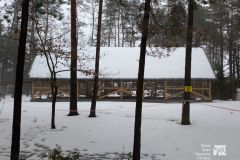 Drewniana konstrukcja podłużnego budynku w trakcie budowy, przed nią kilka drzew, a na jednym z nich tabliczka z napisem „Teren budowy wstęp zabroniony”. Okolica pokryta śniegiem.