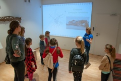 Kobieta opowiadająca i przeprowadzająca dzieci po muzeum w tle filmik od projektora 