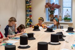 Dzieci siedzą przy dużym stole i wykonują prace plastyczne. Na stole leży kilka czarnych kapeluszy i cylindrów