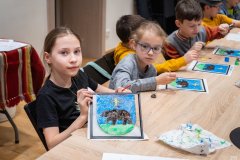 Dzieci siedzą przy stole i wykonują prace plastyczne. Dziwczynka prezentuje gotowy obrazek.