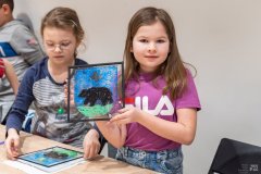 Dwie dziewczynki stoją przy stole. Jedna z nich prezentuje obrazek wykonany na szkle, przedstawiający dawny herb Ostrołęki