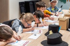 Dzieci siedząc przy stole piszą ptasim piórem na kartach edukacyjnych