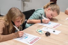 Dwie dziewczynki siedząc przy stole piszą ptasim piórem na kartach edukacyjnych