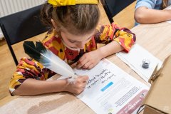 Dziewczynka siedząc przy stole pisze ptasim piórem na karcie edukacyjnej