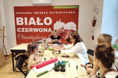 Uczestnicy warsztatów, dzieci i dorośli siedzą przy stołach i wykonują patriotyczne ozdoby. W tle na ścianie duży, biało-czerwony banner z napisem „Biało-czerwona Obowiązkowo!”