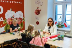 Dwie kobiety i dwie dziewczynki siedzą przy stole i wykonują biało-czerwone ozdoby.