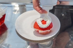 Na białym talerzyku leży czerwone ciastko (babeczka), na które nakładana jest bita śmietana z pojemnika pod ciśnieniem
