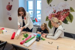 Jedna kobieta stoi przy stole przecinając bibułę, inna siedzi z dziewczynką i razem wykonują ozdoby.