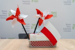 Dwa biało-czerwone wiatraczki i biało-czerwona flaga wetknięte w plastikowy, prostokątny koszyczek. W tle na ściance loga Muzeum Kultury Kurpiowskiej