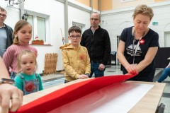 Dorośli i dzieci stoją przy stole, na którym leży duża biało-czerwona. Kobieta demonstruje sposób składania flagi.