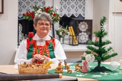 Przy stole siedzi twórczyni w stroju kurpiowski, przed nią stoi mała choinka świąteczna oraz „byśki” i „nowe latka”