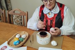 Kobieta w tradycyjnym stroju kurpiowskim , siedząc przy stole wykonuje pisankę wielkanocną. Przed twórczynią stoi zestaw do rozgrzewania wosku, obok którego leży pisanka. Nieco dalej leży koszyczek z pięcioma gotowymi pisankami.