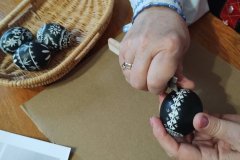 Dłonie wykonujące pisankę wielkanocną. Obok leży gotowa pisanka, nieco dalej koszyk z trzema gotowymi pisankami.