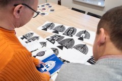 Dwóch mężczyzn ogląda ilustracje wycinanek leżące na stole. Widok z za ramion