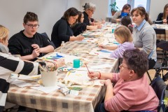 Kilkanaście osób, dorośli i dzieci, siedzą przy długim stole i malują