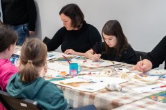 Kilkana osób, dorośli i dzieci, siedzą przy długim stole i malują