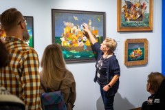 Kilka osób słucha przewodniczki wskazującej dłonią na jeden z obrazów na wystawie