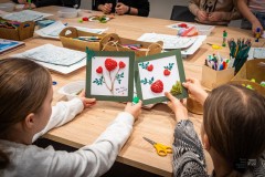 Dłonie dwojga dzieci siedzących przy stole, trzymających dekoracje w ramce jedna obok drugiej