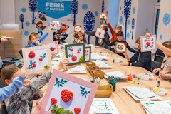 Kilkoro dzieci siedzących przy dużym stole, prezentujących do zdjęcia niewielkie dekoracje w ramkach. W tle, na ścianach zimowe dekoracjez motywem wycinanki kurpiowskiej  i banner z dużym napisem „Ferie w Muzeum”