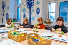 Cztery dziewczynki siedzące przy stole i wykonujące wycinankę. W tle okna, a na ścianie zimowa dekoracja z motywem wycinanki kurpiowskiej