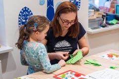 Przy stole siedzi kobieta wykonująca wycinankę. Obok niej stoi dziewczynka i przygląda się, w tle na ścianie dekoracja z motywem wycinanki kurpiowskiej.