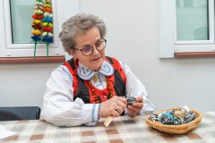 Kobieta w stroju kurpiowskim siedzi przy stole, na którym leży koszyk z pisankami. W tle ściana z oknami. Na parapecie ustawione dwie palemki wielkanocne.