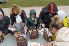 Uczestnicy pikniku Fortach Bema stojąc przy stole przy rozwiązują zadania na kartach edukacyjnych. Na stole znajdują się tekturowe  walizki i materiały edukacyjne