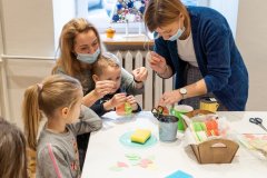 Przy stole dwie kobiety, dziewczynka i chłopiec. Jedna z kobiet trzyma chłopca na kolanach, druga pochyla się na stołem, obydwie trzymają ozdoby choinkowe , tzw. światy wykonane z opłatka.