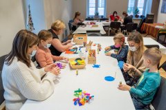 Uczestnicy zajęć siedzą przy stołach, wykonują ozdoby świąteczne. Na stołach wiele materiałów i przyborów do tworzenia. W tle okna.