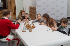 Przy stole siedzą dzieci, kobieta z chłopcem na kolanach oraz mężczyzna w kurpiowskim stroju ludowym. Wykonują pieczywo obrzędowe. Na stole gotowe pieczywo – byśki i nowe latka.