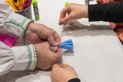 Dłonie dwóch osób na tle stołu. Jedne dłonie trzymają bibułę złożoną w harmonijkę, drugie zawiązują nitkę na bibule