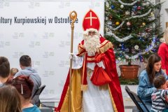 Mężczyzna w stroju Świętego Mikołaja. Po bokach kadru fragmenty stołów, przy których siedzą uczestnicy zajęć. W tle choinka świąteczna oraz ścianka MKK z logotypami