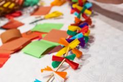 Na stole tradycyjny łańcuch choinkowy z papieru i słomy. Obok łańcucha materiały i przybory do tworzenia.