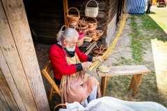 Twórtca w stroju kurpiowskim siedzący przy drewnianym budynku gospodarczym trzyma w słoni pęk wikliny. Za nim, na stoliku stoi kilka wyplecionych koszyków