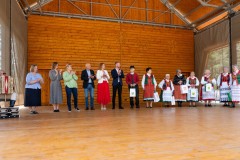 Na scenie stoi kilkanaście osób, kila jest w tradycyjnym stroju kurpiowskim. Mężczyzna z lewej strony siedzi grając na harmonii.