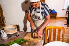 Kobieta w fartuchu z motywem wycinanki kurpiowskiej i chustce na głowie, kroi chleb.