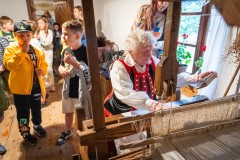 W kurpiowskiej izbie kilkoro dzieci przygląda się jak kobieta wykonuje tkaninę na krośnie