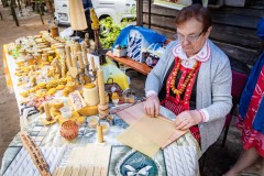 Kobieta w stroju kurpiowskim siedzi przy stole i wykonuje świecę z wosku. Na stole wiele wyrobów woskowych.