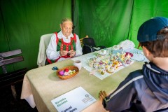 Kobieta w stoju kurpiowskim siedząc przy stole wykonuje koronkę, obok niej na białej serwecie ułożone wyroby koronkarskie. Jedna osoba przygląda się jej pracy.