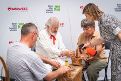 Mężczyzna w stroju kurpiowskim uczy trzy osoby wyplatania z wikliny