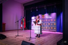 Na scenie kobieta w stroju kurpiowskim przemawia do mikrofonu. W tle ścianki z motywami gminy Kadzidło.