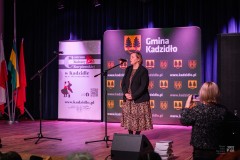 Na scenie kobieta przemawia do mikrofonu. W tle ścianki z motywami gminy Kadzidło. Przed sceną ktoś robi zdjęcia smartfonem.