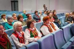 Ponad dwadzieścia osób siedzi na widowni. W oddali stoi kobieta z aparatem fotograficznym.