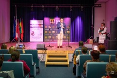 Na scenie mężczyzna przemawia do mikrofonu. W tle ścianki z motywami gminy Kadzidło. Z prawej strony, na scenie, stoi kobieta w stroju kurpiowskim