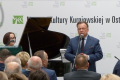Mężczyzna stojący przy mikrofonie wraz z kobietą przed widownią na tle napis MKK i fortepian